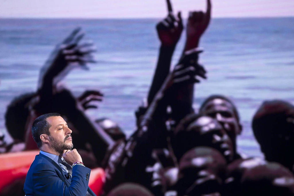 Matteo Salvini, ministro do Interior italiano, discursa em frente a painel com foto de imigrantes africanos em barco no Mediterrâneo